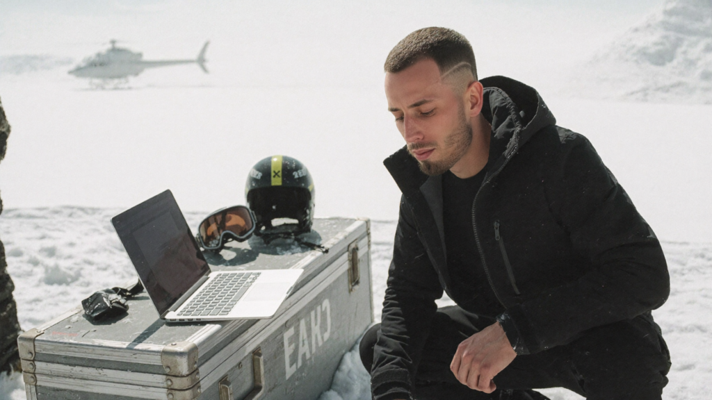 A man crouched over a laptop on an equipment case on a glacier snowfield in flat high-key overcast light