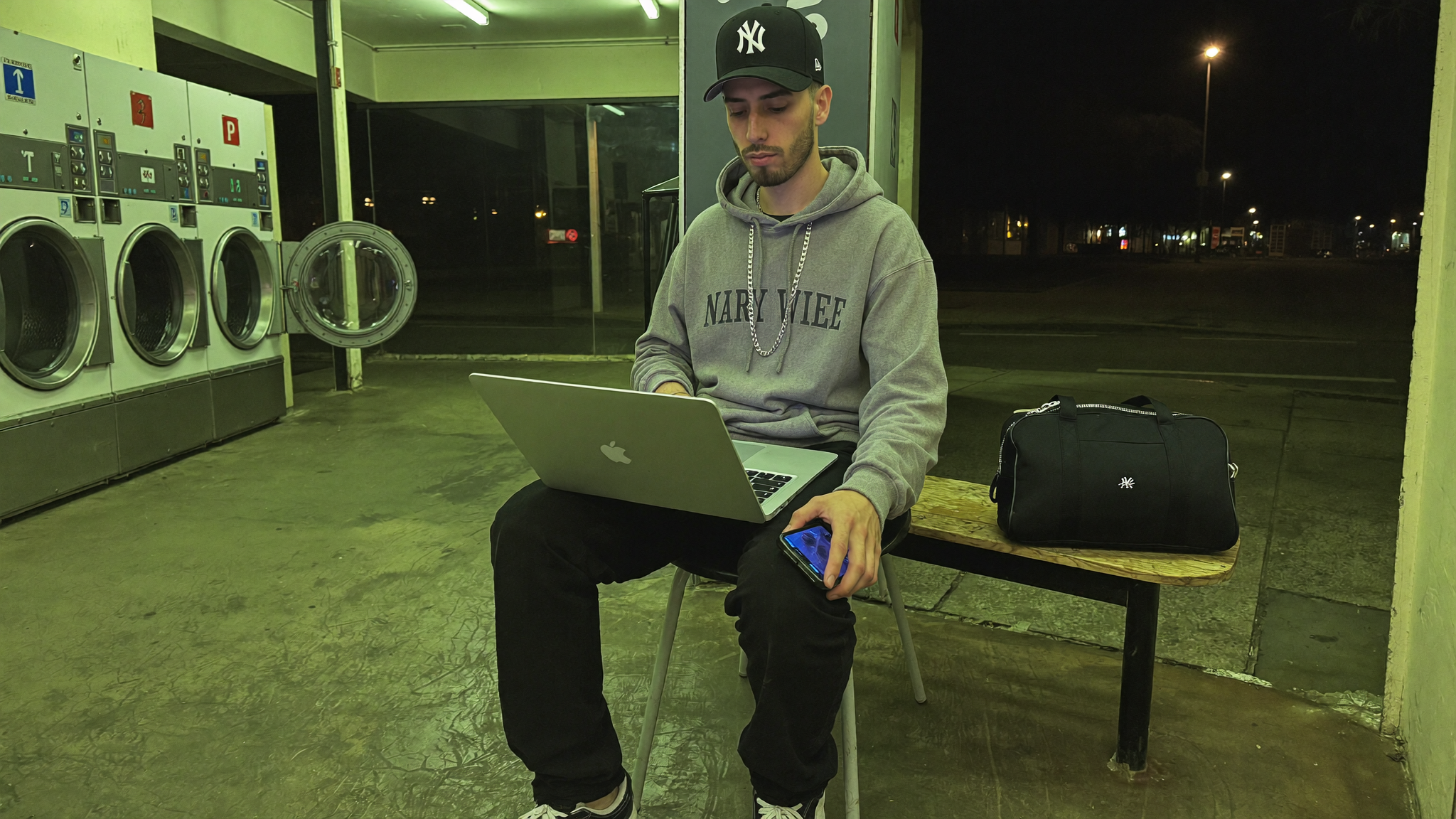 A man working on a laptop in an empty coin laundromat at two in the morning under flat green fluorescent light