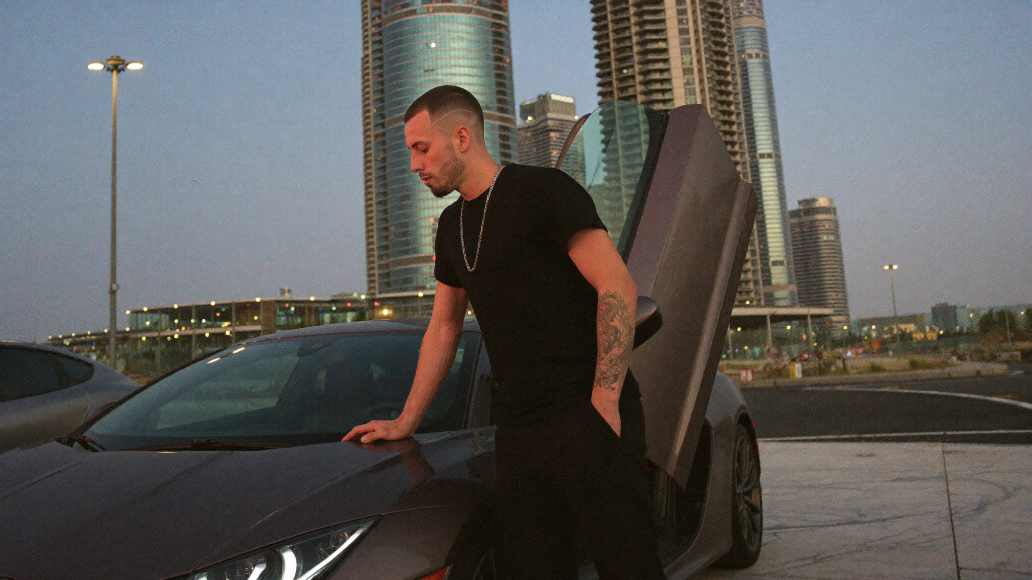 Founder leaning against a matte grey supercar with the door raised on a marble forecourt in Dubai at dusk