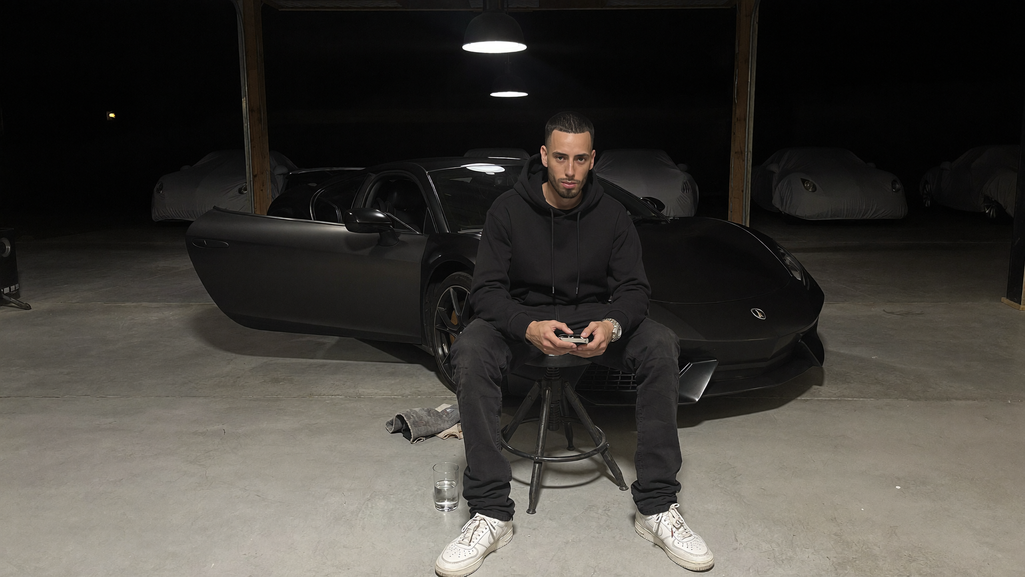 Cody Wise seated on a workshop stool in front of a matte supercar in a dark private garage
