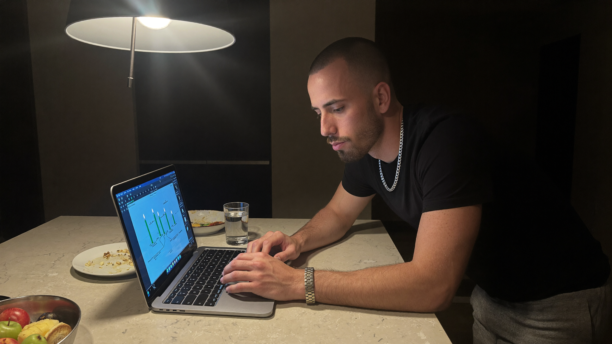 A founder checking a candlestick chart on a laptop at a kitchen island after midnight