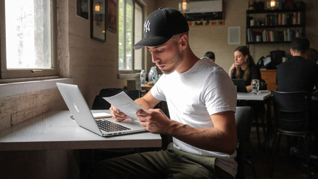 Laptop and printed documents on a coffee shop table by a window, representing reviewing Google Ads campaign performance against real sales data